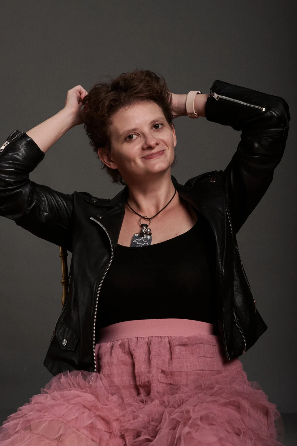 Kate in a leather jacket and pink skirt, smiling with her hands raised to her hair